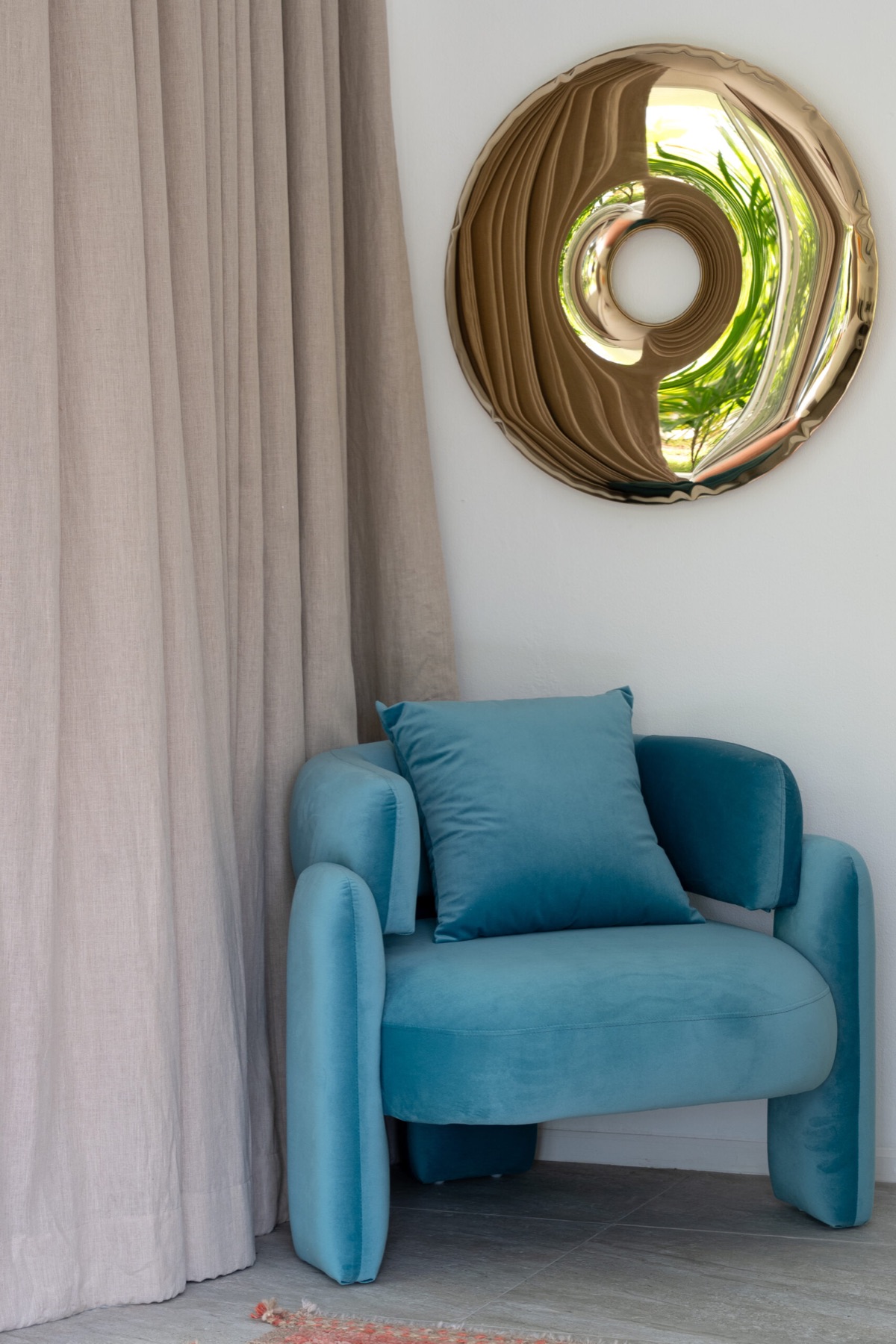 Velvet armchair in teal beside linen curtains and a bronze mirror